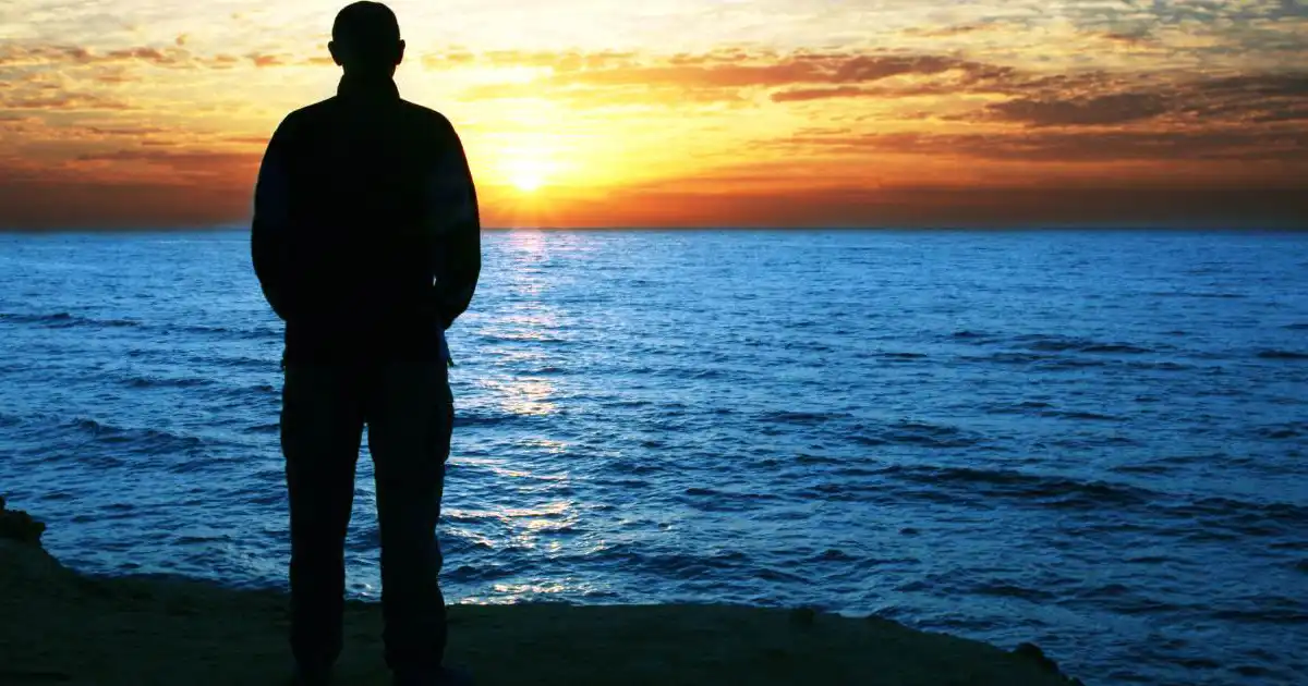 Person standing quietly at the edge of the sea during sunrise, facing the horizon in silent reflection